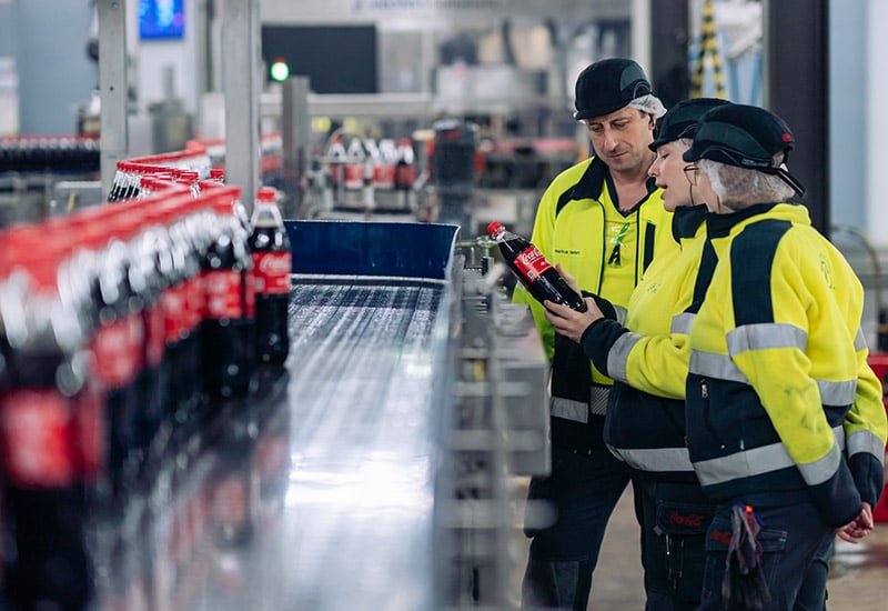 Coca-Cola Werk in Mönchengladbach mit 3 Mitarbeitenden vor dem Produktionsband